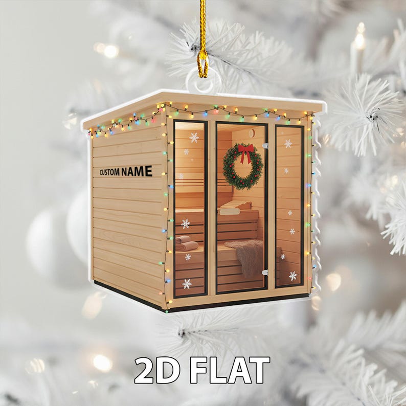 May include: A wooden sauna Christmas tree ornament, decorated with a wreath and fairy lights. The ornament has the text "CUSTOM NAME" and "2D FLAT". The sauna features glass doors and windows, and a gold hanging string.