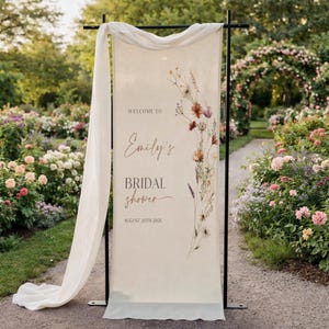 May include: A cream-colored bridal shower welcome sign with floral accents and the text "Welcome to Emily's Bridal Shower August 20th 2026." The sign is draped with a white fabric and set on a black metal frame, with a garden background.