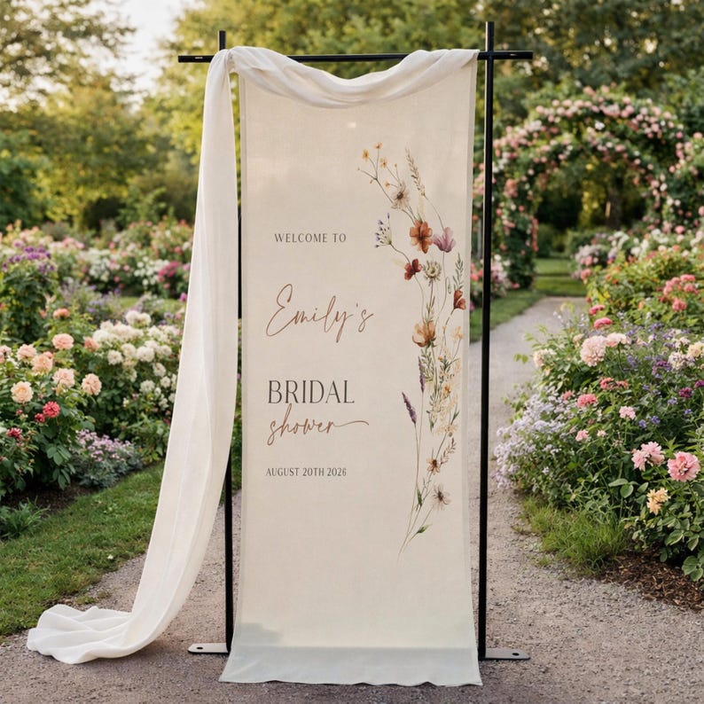May include: A cream-colored bridal shower welcome sign with floral accents and the text "Welcome to Emily's Bridal Shower August 20th 2026." The sign is draped with a white fabric and set on a black metal frame, with a garden background.