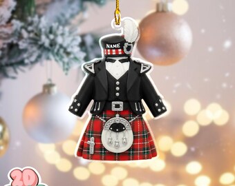 May include: A Scottish kilt Christmas tree ornament. It features a black jacket, a red and green plaid kilt, and a white feather on the hat. The ornament has a gold hanging string and the word "NAME" is printed on the hat. Silver accents complete the design.