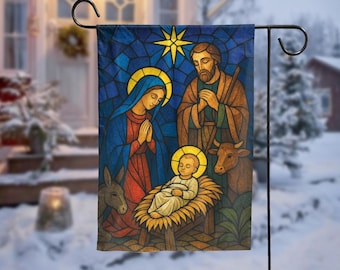 Può includere: Una bandiera da giardino in stile vetrata raffigura la scena della Natività. La bandiera presenta Maria, Giuseppe e il bambino Gesù, con una stella, un asino e una mucca. La bandiera è esposta su un paletto da giardino in metallo nero in un ambiente esterno innevato.