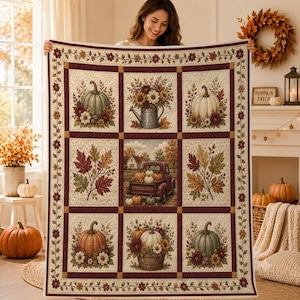 Op de afbeelding: Een rechthoekige quilt met een raster van negen panelen met herfstthema-illustraties, waaronder pompoenen, zonnebloemen, herfstbladeren, een metalen gieter en een rode pick-up truck vol pompoenen, omrand door een bloemmotief.