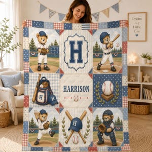 May include: A quilted blanket featuring a baseball theme with illustrations of teddy bears in uniforms, baseballs, bats, and a glove. The design includes the letter H, the name Harrison, and a red, white, and blue colour palette with plaid borders.