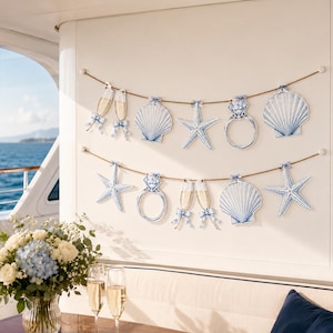 May include: Two horizontal twine garlands featuring hanging cutouts of blue and white seashells, starfish, diamond rings, and champagne flutes. Each cutout is decorated with a small blue ribbon bow at the top, creating a nautical themed display.
