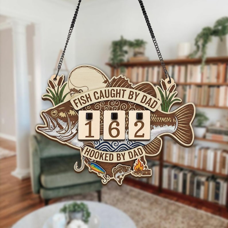 May include: Wooden sign with a fish design, the text "FISH CAUGHT BY DAD" and "HOOKED BY DAD". The sign has a chain for hanging and features a counter displaying the number 162. Decorative elements include fishing lures and a campfire.