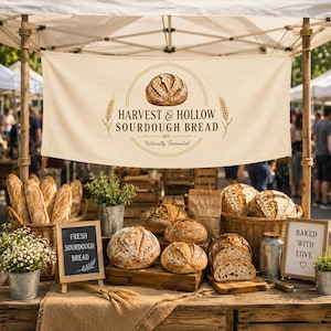 May include: A cream-colored fabric banner with the text Harvest & Hollow Sourdough Bread and a bread illustration hangs above a market stall display featuring various round sourdough loaves in wicker baskets and on wooden crates.
