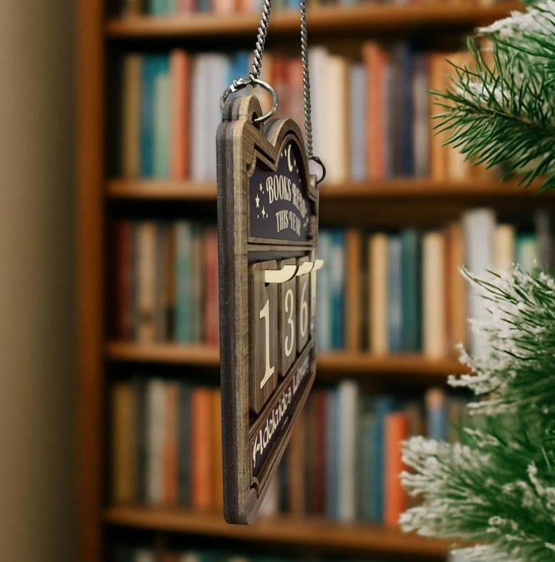 May include: Wooden sign with the text "BOOKS READ THIS YEAR" and the number "136". The sign is hanging from a silver chain. The background is a bookshelf.