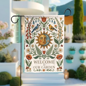 Może przedstawiać: Flaga ogrodowa z powitalnym przesłaniem. Flaga przedstawia wzór słońca i księżyca z ptakami, kwiatami i napisem "WELCOME to OUR GARDEN". Flaga jest prezentowana na czarnym metalowym stojaku, idealna do dekoracji zewnętrznych.