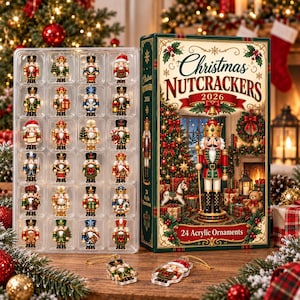 Op de afbeelding: Een set van 24 acryl notenkraker ornamenten in een doorzichtige plastic tray naast een rechthoekige doos met het opschrift Christmas Nutcrackers 2026. De doos toont een illustratie van een notenkraker, een kerstboom en cadeaus.
