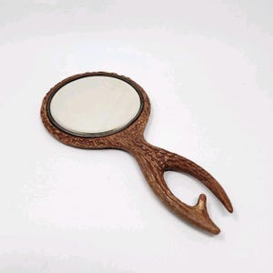 May include: A brown hand mirror with an oval mirror and a handle designed to resemble a deer antler. The mirror frame is a darker brown, and the handle has a textured, naturalistic appearance.