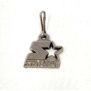 May include: Silver-colored metal zipper pull with the Starter logo. The logo features a stylized "S" shape, a star, and the word "STARTER" in capital letters. The pull has a small clasp at the top for attachment.
