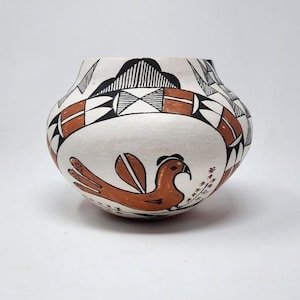 May include: A white ceramic pot with a rounded shape, featuring a brown bird design and geometric patterns in black and brown. The pot has a wide opening and is decorated with traditional Native American motifs.