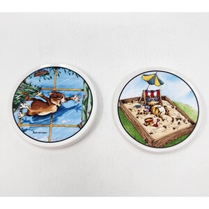 May include: Two round ceramic coasters with cat illustrations. One shows a brown cat reaching out a window. The other depicts a cat in a sandbox with sunglasses, a beach chair, and an umbrella. Both have white borders.