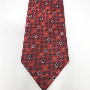 May include: A patterned necktie in shades of red, orange, and brown. The tie features a repeating diamond pattern with small, oval-shaped accents. The tie is made of a smooth fabric and is suitable for formal or business attire.