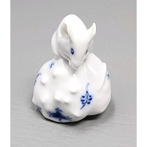 May include: A white porcelain figurine of a curled-up animal, possibly a mouse or a rabbit. The figurine has blue accents and floral patterns. The animal's head is tilted downward, and its body is in a curled position. The figurine is on a gray background.