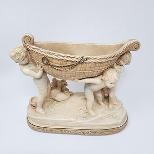 May include: A beige ceramic planter featuring three cherubic figures supporting a woven basket. A duck figurine rests on the base. The planter has a decorative base and is suitable for small plants or flowers.