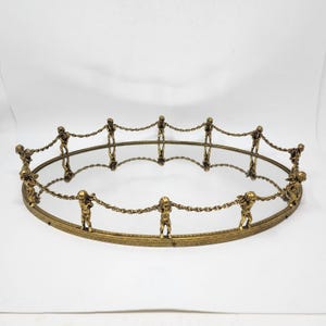 May include: An antique gold-toned decorative tray with a mirrored surface. The tray is oval-shaped and features small figures holding chains around the perimeter. The figures are connected by chains, creating a unique and ornate design.