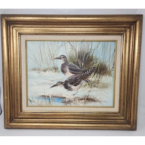 May include: An oil painting of two birds in a marshy landscape, framed in a gold-colored, ornate wooden frame. The birds have brown and white speckled feathers, long beaks, and are standing near a small pool of water. The background features tall, dried grasses.