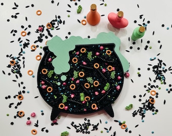 Pode incluir: Uma bandeja preta em forma de caldeirão cheia de granulado colorido, rodeada por pequenas garrafas com rolhas. O caldeirão tem um topo verde e borbulhante e granulado em forma de anel laranja. A cena é festiva e caprichosa, perfeita para o Halloween.