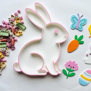 Puede incluir: Una bandeja blanca con forma de conejo con borde rosa, rodeada de decoraciones temáticas de Pascua. Incluye una mariposa, un pollito, una zanahoria, una flor, un conejo y un huevo. También pasta de varias formas y colores.