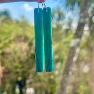 May include: A pair of teal rectangular earrings with silver hooks. The earrings have dark blue veining. The earrings are displayed against a backdrop of a blue sky and green foliage.