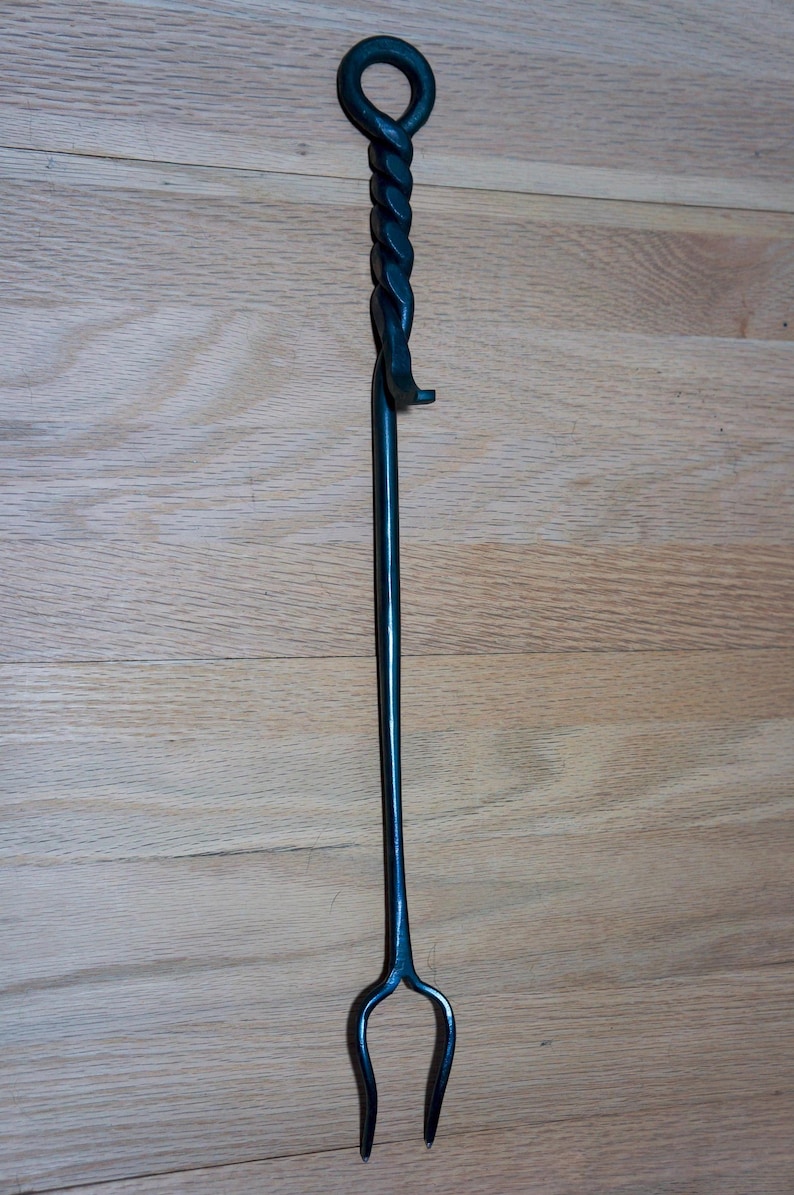 May include: A hand-forged, dark metal fireplace poker. The poker features a twisted handle with a loop at the top and a forked end. The tool is designed for tending to a fire and is shown against a wood background.