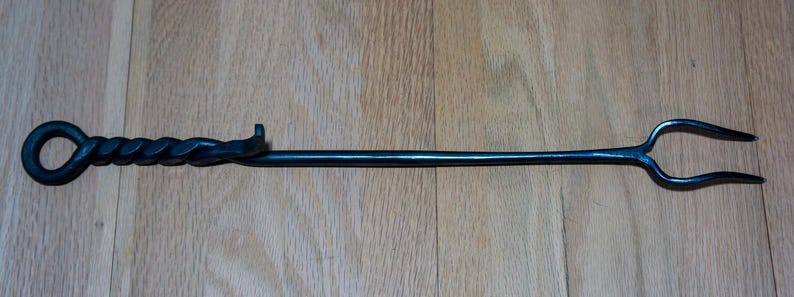 May include: A black, hand-forged fireplace poker with a twisted handle and a loop at the end. The poker measures approximately 30 inches in length and has a two-pronged fork at the opposite end.