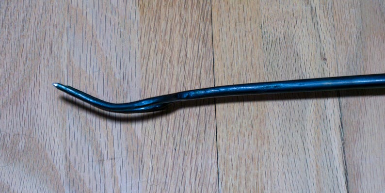 May include: A dark blue metal pry bar with a curved, pointed end. The tool is lying on a wooden surface. The pry bar is designed for leverage and prying objects apart.