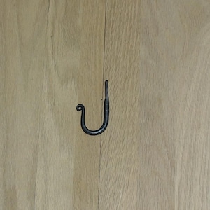 May include: A black, wrought iron hook with a curved design. The hook is mounted on a light-colored wooden surface. The hook has a decorative, curled end and a simple, functional design.