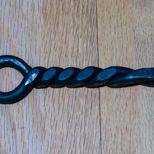 May include: A black, hand-forged fireplace poker with a twisted handle and a loop at the end. The poker is made of iron and sits on a wooden surface.