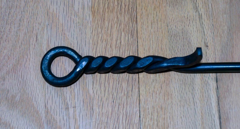 May include: A black, hand-forged fireplace poker with a twisted handle and a loop at the end. The poker is made of iron and sits on a wooden surface.