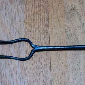 May include: A black, forged metal fire poker with a two-pronged fork at one end. The tool has a long, slender handle and is designed for use in a fireplace or outdoor fire pit. The metal has a slightly textured surface.