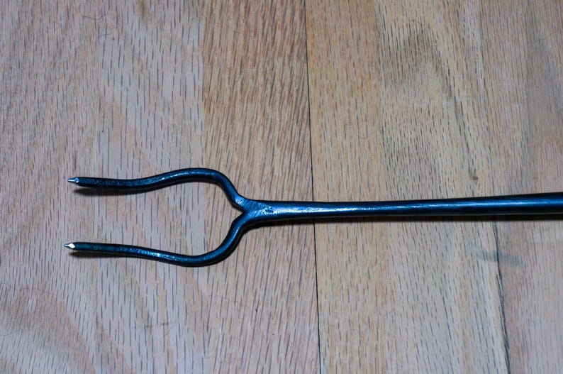 May include: A black, forged metal fire poker with a two-pronged fork at one end. The tool has a long, slender handle and is designed for use in a fireplace or outdoor fire pit. The metal has a slightly textured surface.