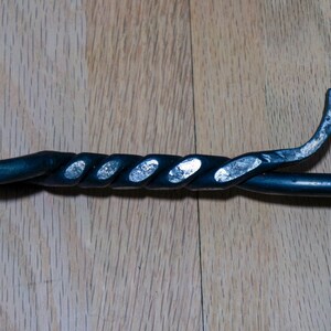 May include: A black metal fireplace poker with a twisted handle and a curved end. The handle has a decorative, spiral design. The poker is set against a wooden background.