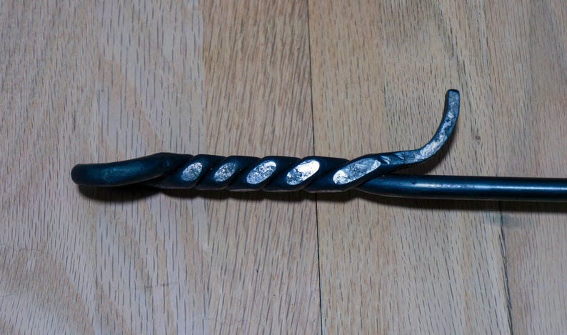 May include: A black metal fireplace poker with a twisted handle and a curved end. The handle has a decorative, spiral design. The poker is set against a wooden background.