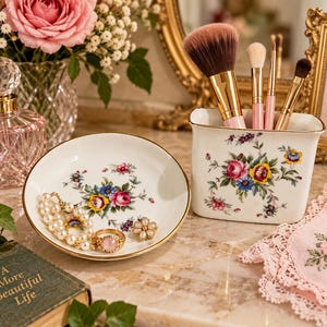 May include: A white porcelain vanity set featuring a round trinket dish and a rectangular brush holder, both decorated with colorful floral patterns and gold trim. The dish contains a pearl bracelet, a gold ring, and a floral stud earring.