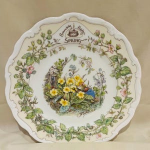 Puede incluir: Plato decorativo crema con borde festoneado, que presenta una escena primaveral con un ratón, flores y el texto "Brambly Hedge SPRING". El plato tiene un borde floral y una ilustración central.