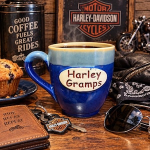 May include: A two-toned blue ceramic mug featuring a cream-colored oval plaque with the text Harley Gramps embossed on the front. The mug has a glossy finish with a lighter blue rim and a curved handle, designed for holding hot beverages.