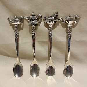 May include: Four silver-toned metal spoons arranged in a row. Each spoon features a unique decorative handle top shaped like a different teapot or oil lamp design. The stems of the spoons are adorned with a small, clear, circular inset stone.