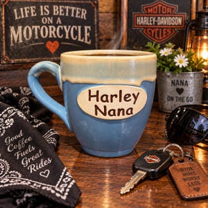 May include: A ceramic mug with a blue base and a cream-colored rim, featuring an oval plaque with the text Harley Nana. The mug has a curved handle and a smooth finish, presented as a functional drinkware item for holding hot beverages.