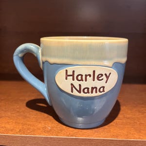 May include: A ceramic mug with a blue base and a light beige rim. The mug has a curved handle and an oval-shaped label with the words "Harley Nana" in brown lettering.