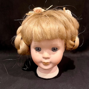 May include: A porcelain doll head featuring blonde hair styled with bangs and side braids, accented by two small pale pink fabric flowers. The doll has blue eyes, painted eyelashes, and a neutral facial expression with a light skin tone.