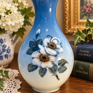 May include: A bulbous ceramic vase featuring a gradient color scheme that transitions from a deep blue at the flared rim to a soft cream at the base. The front is decorated with a hand-painted floral motif of white blossoms with brown centers.