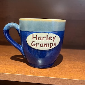 May include: A ceramic mug with a blue and light blue glaze, featuring the text "Harley Gramps" on a white oval. The mug has a curved handle and is designed for hot beverages.
