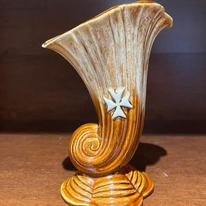 May include: A ceramic vase in a horn shape, with a brown and tan colour scheme and a glossy finish. The vase has a decorative white cross emblem and a curled base. The base is a leaf-like design.
