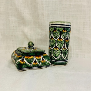 May include: A decorative ceramic box with a lid and a matching cylindrical vase. Both items are hand-painted with a green, yellow, and white floral pattern. The box is square with rounded edges, and the vase is tall and slender.