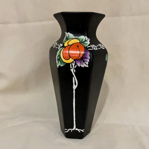 May include: Black vase with a geometric shape and a floral design. The vase features a colorful illustration of a flower with orange, yellow, and purple accents. A white stem and leaves are also present.
