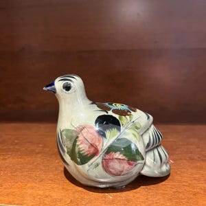 May include: A ceramic bird ornament with a cream-coloured body, featuring painted details. The bird has a black and white striped tail, a blue beak, and is adorned with floral and leaf patterns in shades of red, green, and brown. The ornament is a decorative item.