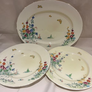 Könnte beinhalten: Ein Set aus drei cremefarbenen Keramiktellern mit Blumen- und Schmetterlingsdesign. Das Set umfasst eine ovale Platte und zwei runde Teller, die mit bunten Blumen und Schmetterlingen verziert sind. Die Teller haben eine zarte, Vintage-Ästhetik.