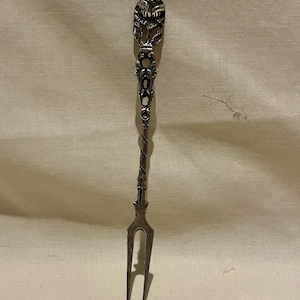 May include: A silver-toned metal cocktail fork featuring an ornate handle with a sculpted, rounded top element and intricate decorative detailing along the stem. The fork has two long, slender tines and a twisted metal design near the base of the handle.
